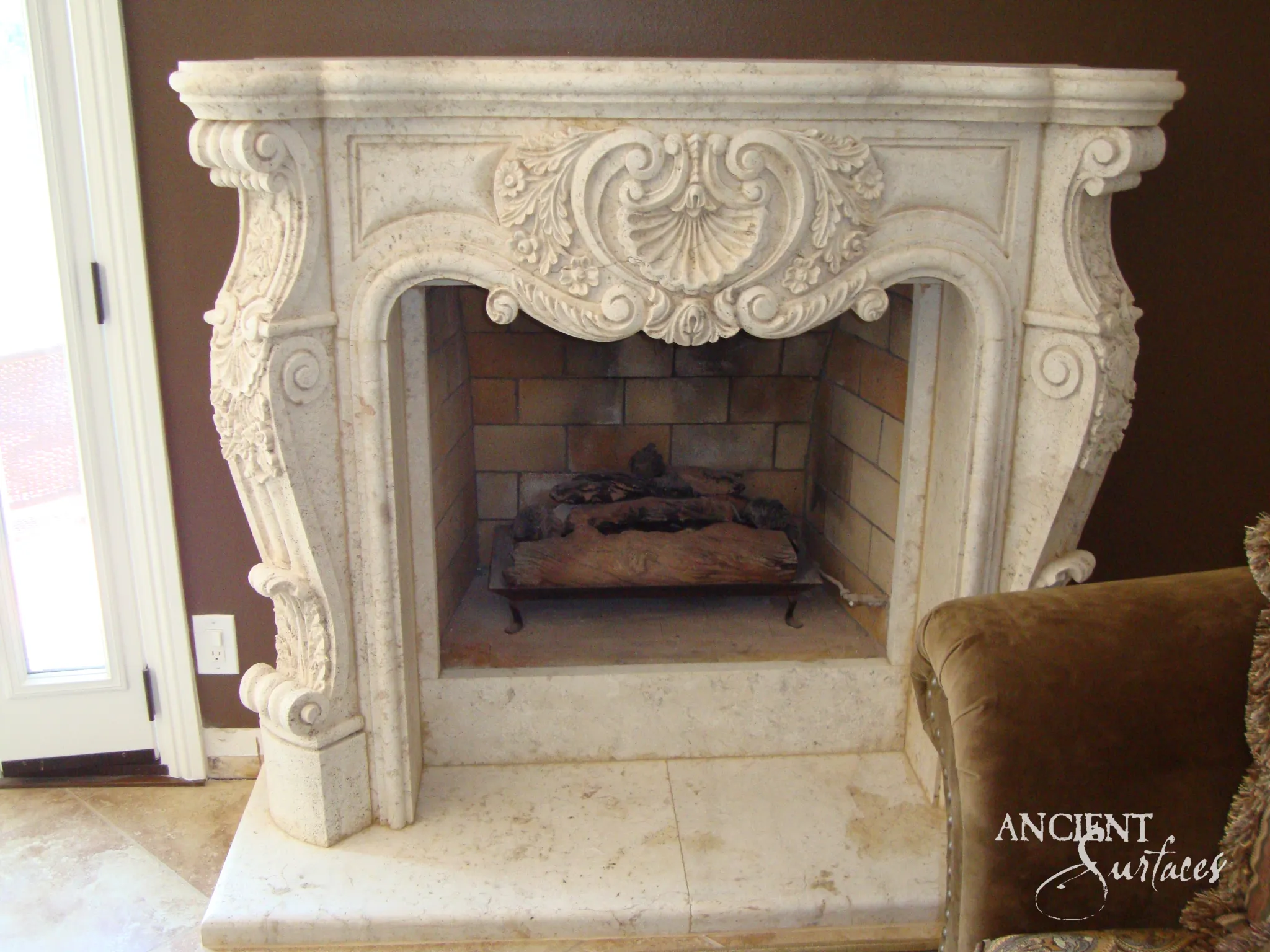 Reclaimed antique limestone mantel from Ancient Surfaces installed against a dark wall, with a serpentine moulded shelf, a carved shell and scroll cartouche on the frieze and volute legs beside a brick firebox