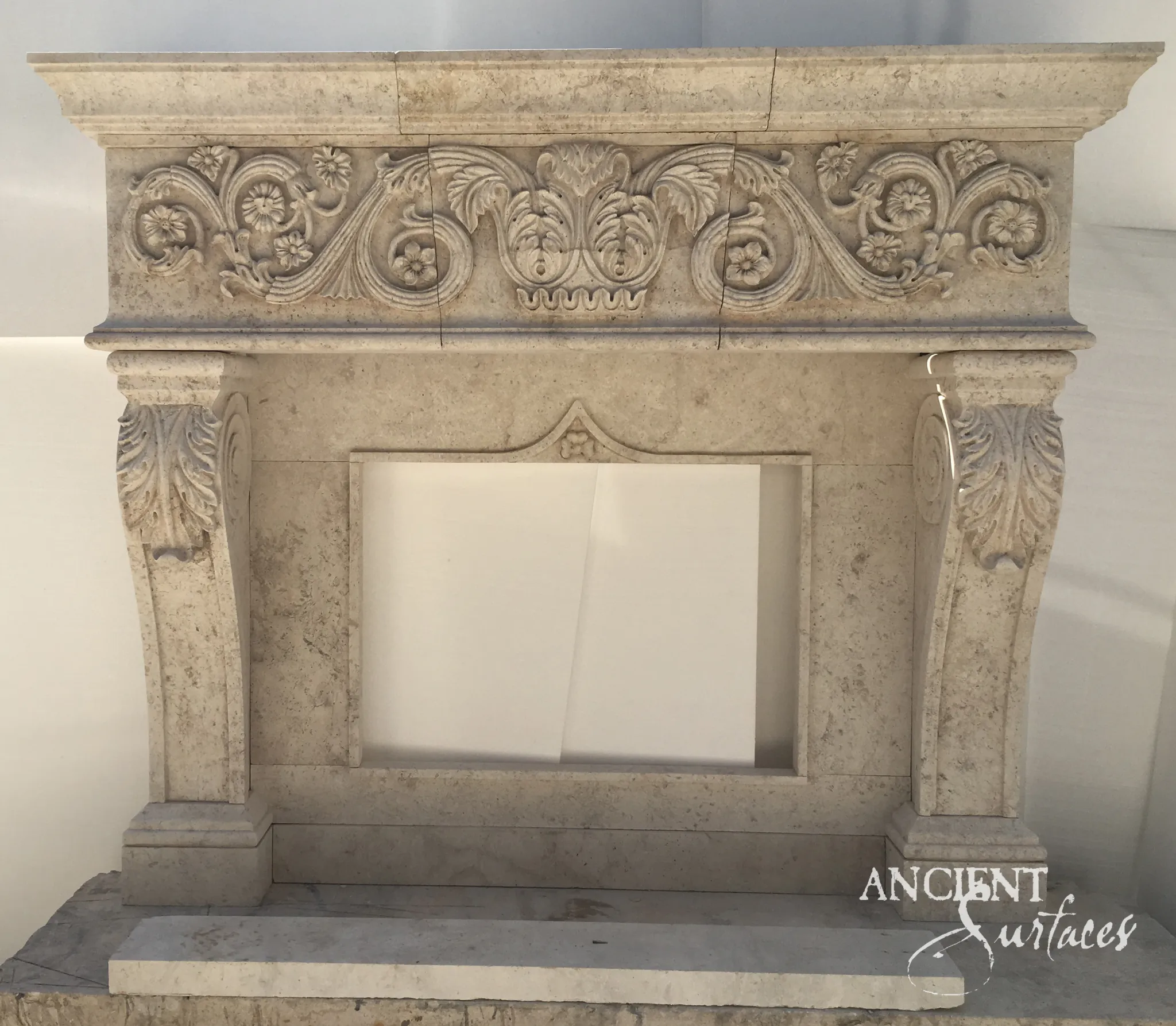 Reclaimed antique limestone mantel from Ancient Surfaces standing before installation, its three block frieze carved with scrolling stems and open flowers around a central palmette, with acanthus corbels above tapering legs