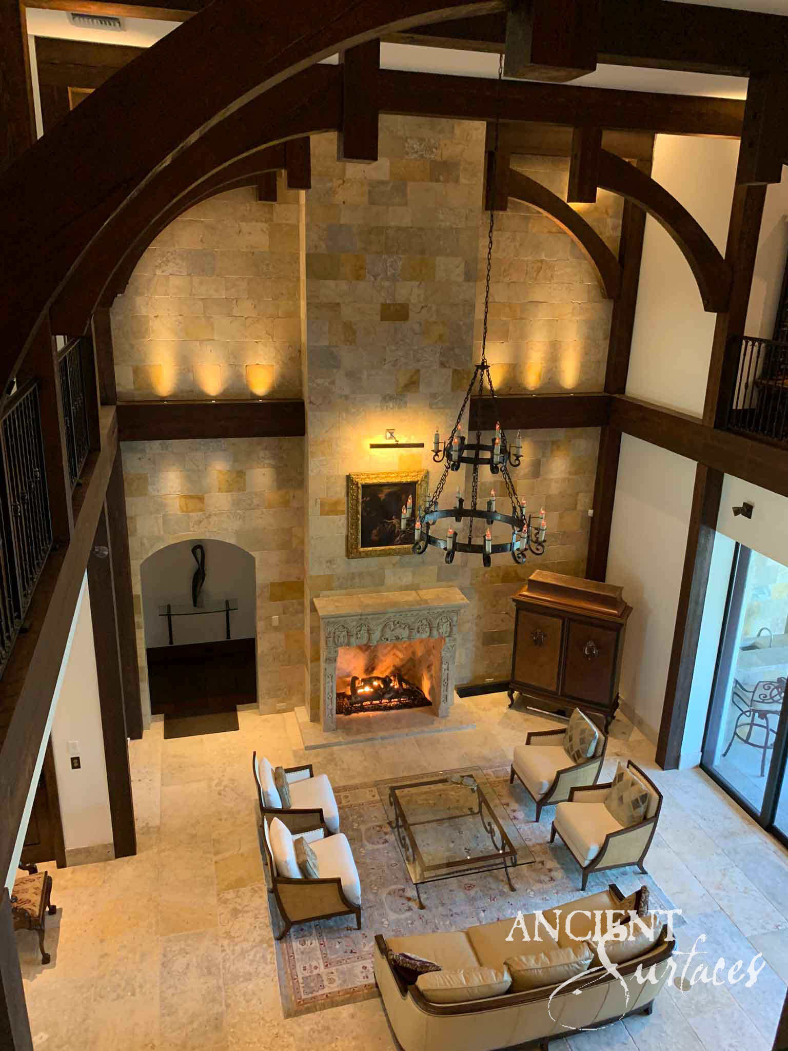 Reclaimed antique limestone mantel from Ancient Surfaces seen from a gallery above, its carved frieze and moulded shelf set into a limestone chimney breast in a double height room under dark timber trusses