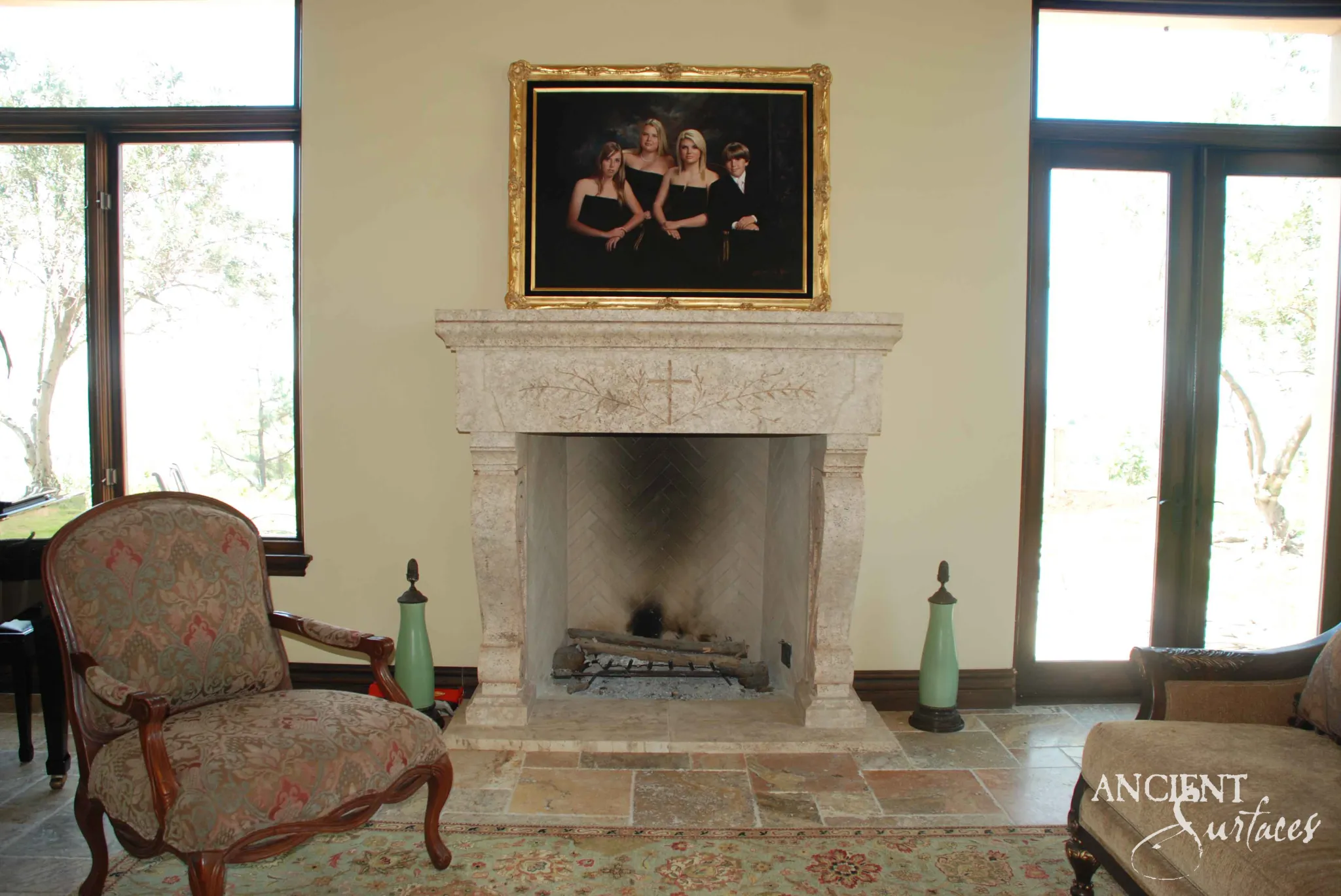 Reclaimed antique limestone mantel from Ancient Surfaces with a flat shelf and a lightly carved frieze of a slim cross with trailing leafed branches, above a herringbone lined firebox between tall windows