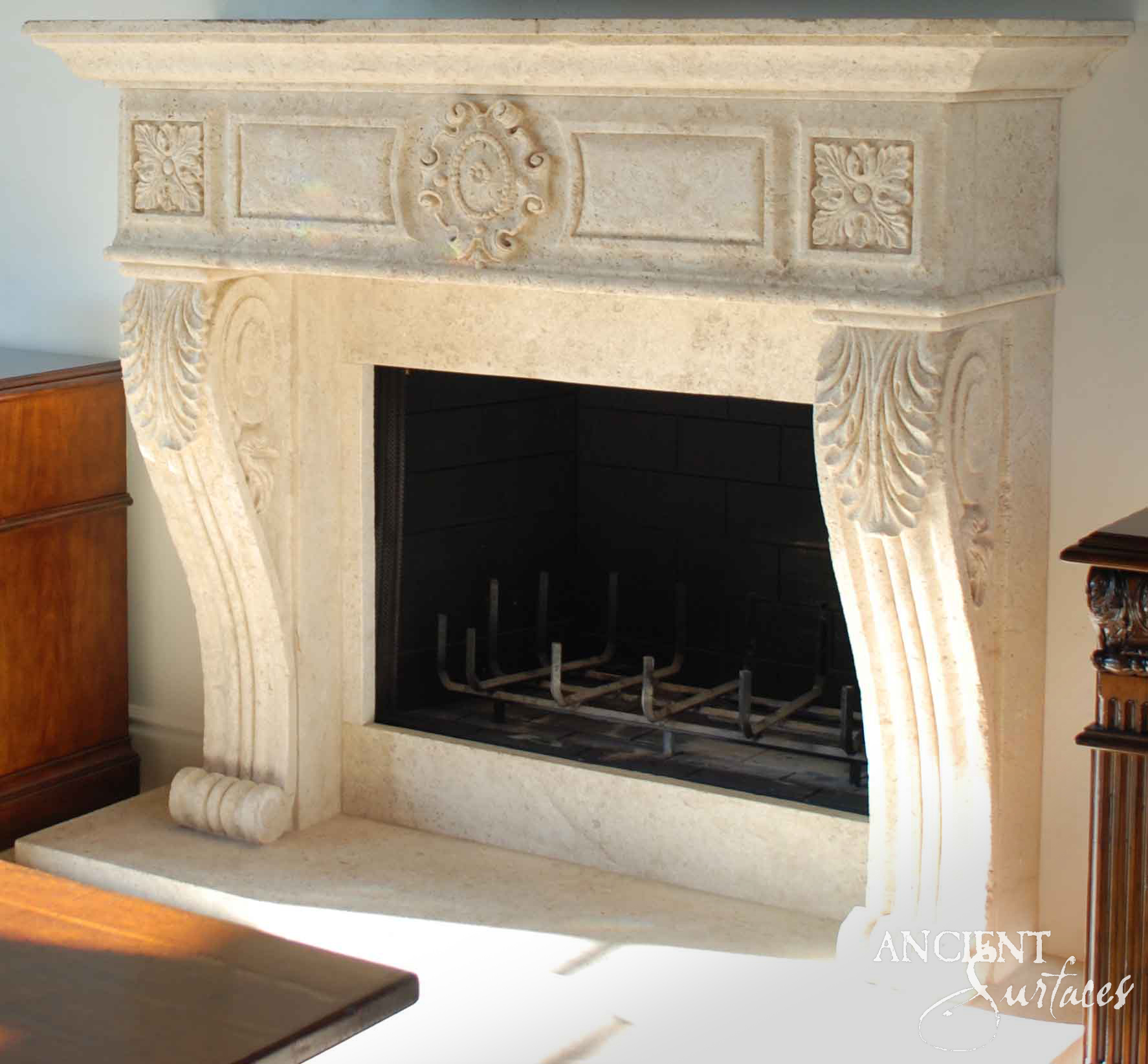 Reclaimed antique limestone mantel from Ancient Surfaces in bright sunlight, its frieze carved with a scrolled oval cartouche between recessed panels and rosette end blocks, above acanthus console legs with volute feet
