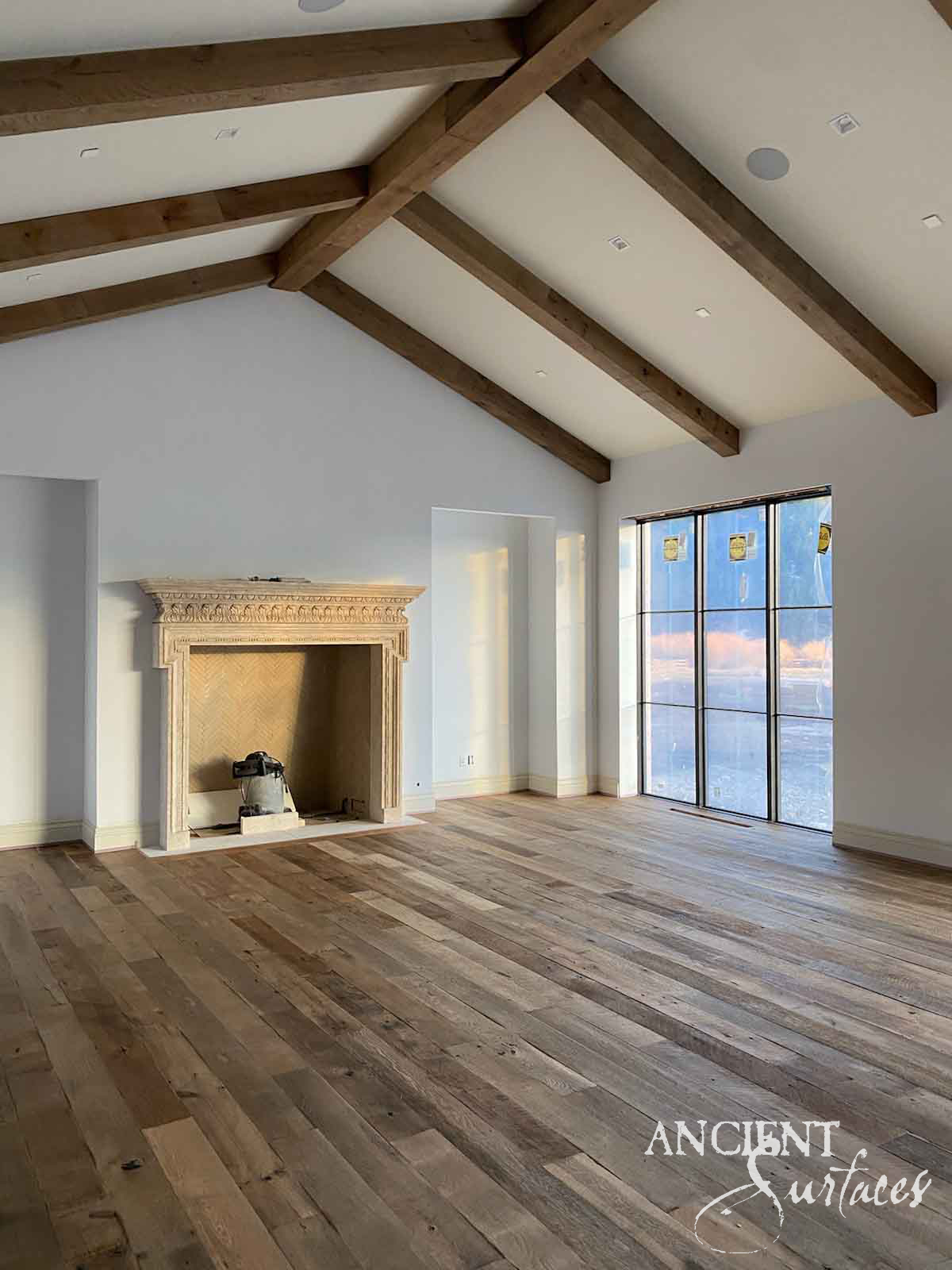Reclaimed antique limestone mantel from Ancient Surfaces in an empty beamed room, its frieze carved with a repeating arcade of scalloped lunettes under a moulded shelf, above a herringbone lined firebox