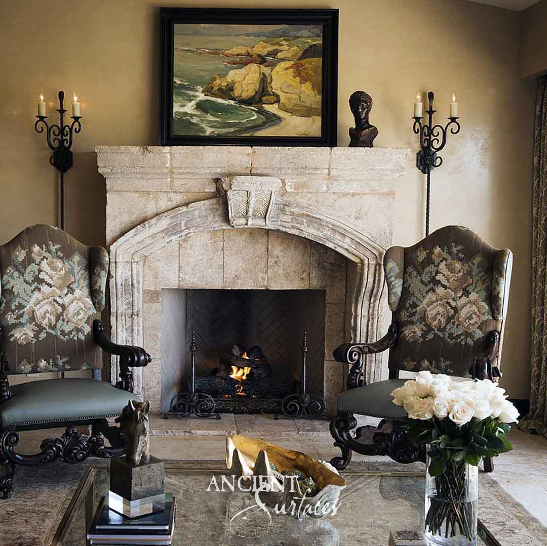 Reclaimed antique limestone mantel from Ancient Surfaces with a broad flattened arch of shaped voussoirs, a carved leaf keystone and a heavy jointed shelf, above a lit herringbone firebox between two upholstered wing chairs