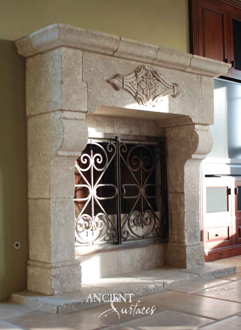 Reclaimed antique limestone mantel from Ancient Surfaces at an angle, with a rounded jointed shelf, a single carved cartouche on a plain frieze, a projecting stone hood over the opening and scrolled iron fire doors