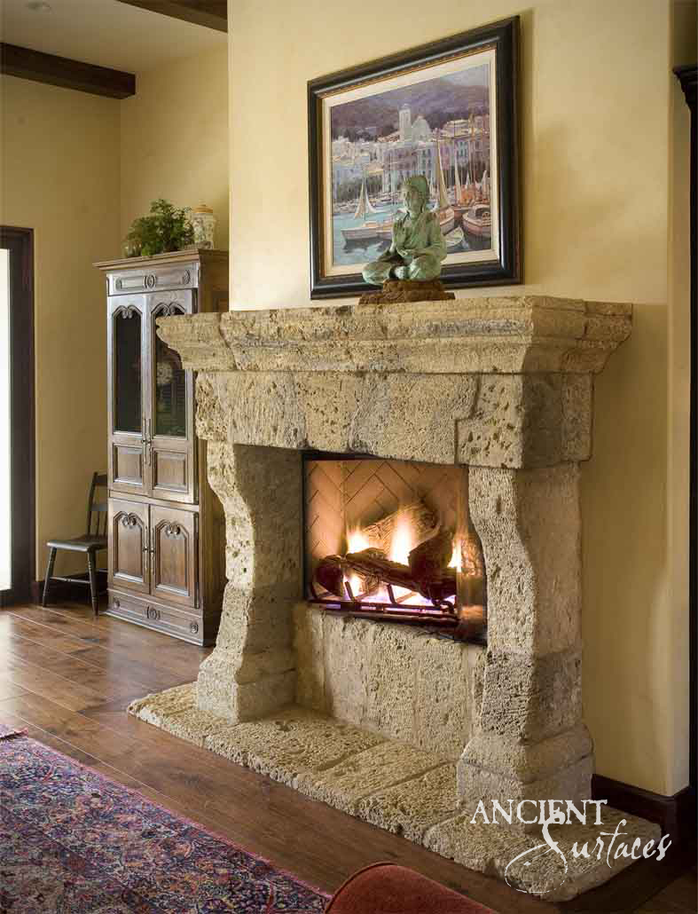 Reclaimed antique limestone mantel from Ancient Surfaces built from rough eroded blocks with a corbelled shelf and thick tapering legs, above a lit herringbone firebox and a rough stone hearth slab